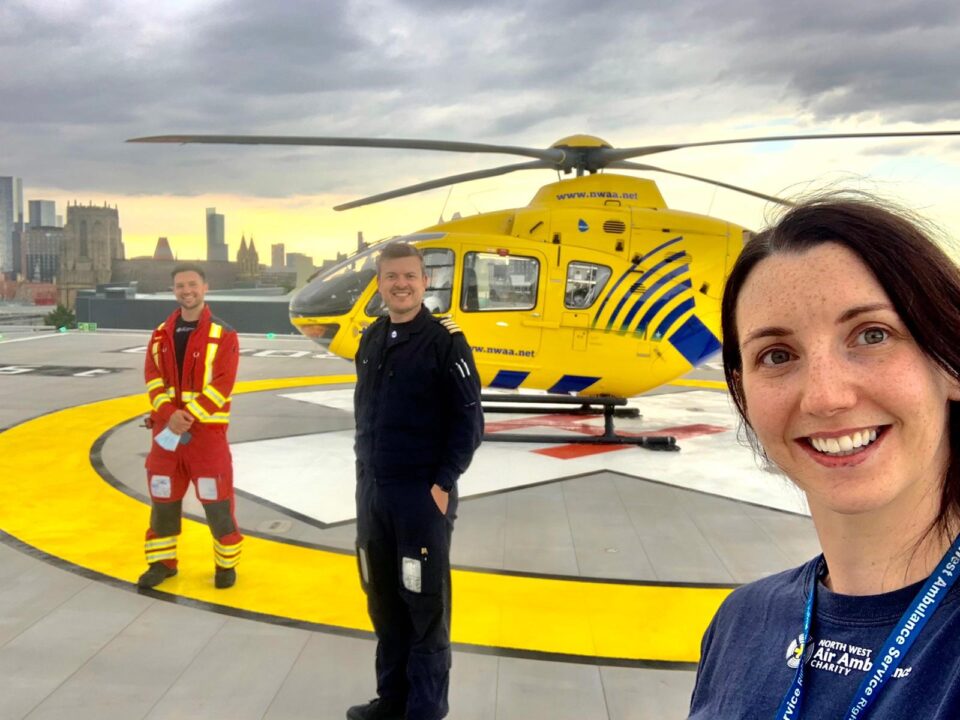 three crew members smiling standing on top of a helipad with helicopter in the background