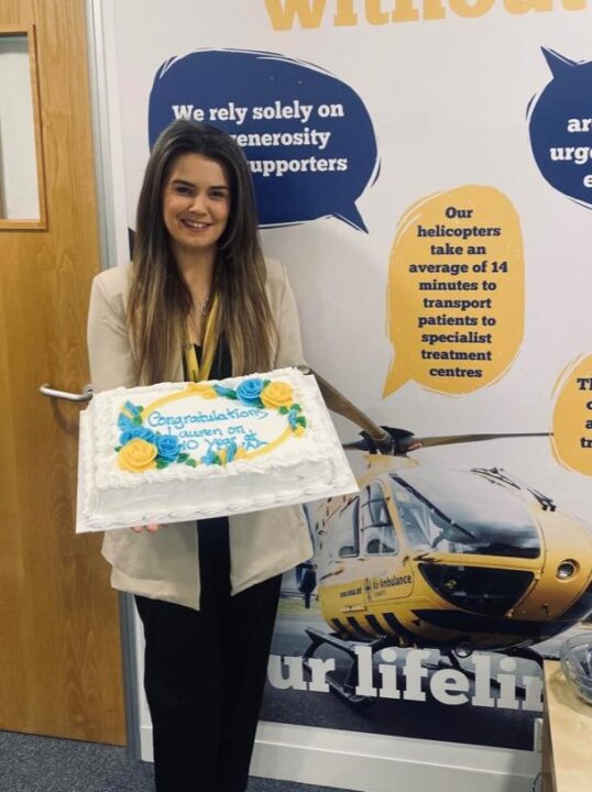 Supporter Engagement Assistant Lauren celebrating her 10 year anniversary working with NWAA. Lauren is holding a white cake with yellow and blue flowers and 'Congratulations' is written on it.