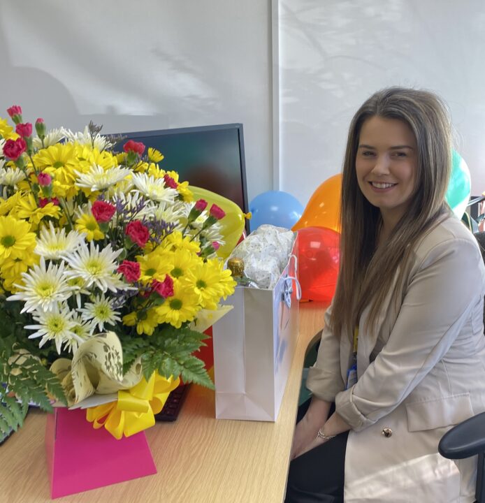 Supporter Engagement Assistant Lauren at her desk with yellow, pink and white flowers, a gift bag and balloons.