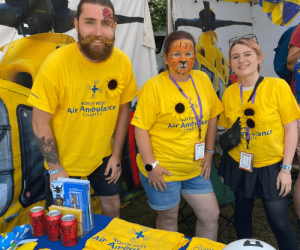 Image of three volunteers. Young man has a beard with glitter on his face and is wearing a yellow t-shirt with North West Air Ambulance Charity written on it. Women stands next to him with tiger face paint (orange face with black lines, black nose and mouth outline) also in a yellow t-shirt with North West Air Ambulance Charity written on it. Young women on the right also in a yellow t-shirt with North West Air Ambulance Charity written on it. All are smiling. 