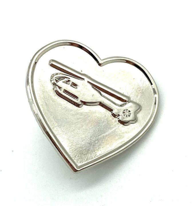 A silver metal heart pin bade with a debossed North West Air Ambulance helicopter in the centre.