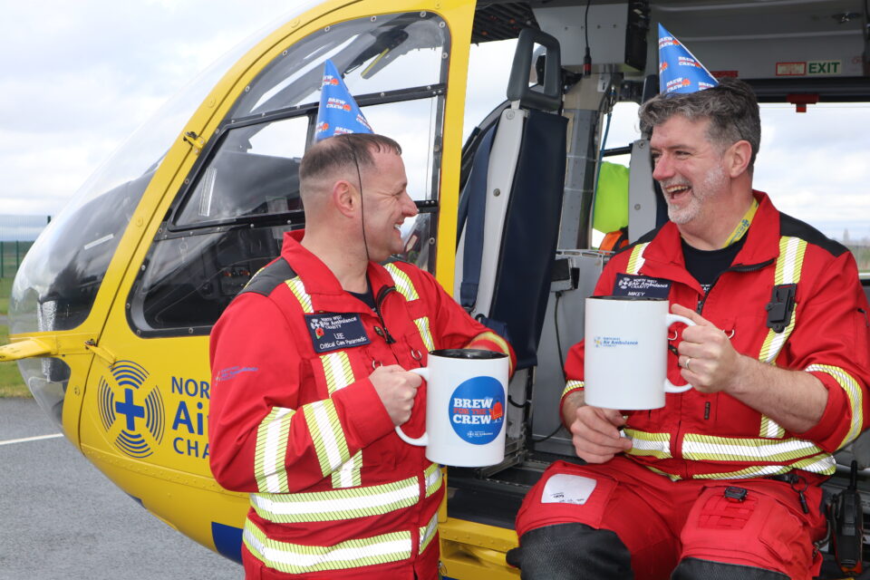 Two male crew members, in red flight suits, sitting on the helicopter, looking at each other laughing. Both have a party hat on and are holding over sized mugs.