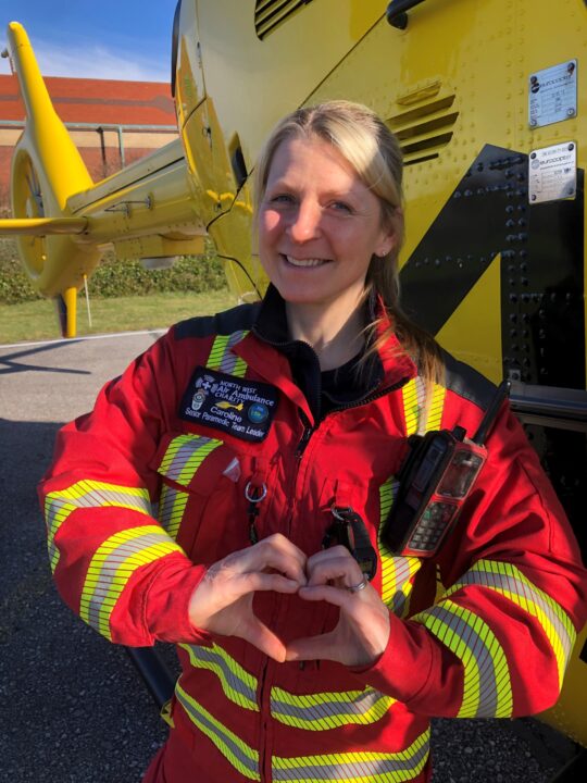 Critical care paramedic in a red flight suit stood in front of a yellow helicopter 