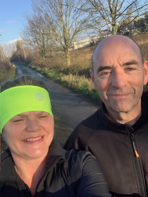 Image of Gary and his wife, who is wear a neon green head-warmer, in running clothes on a country road. 