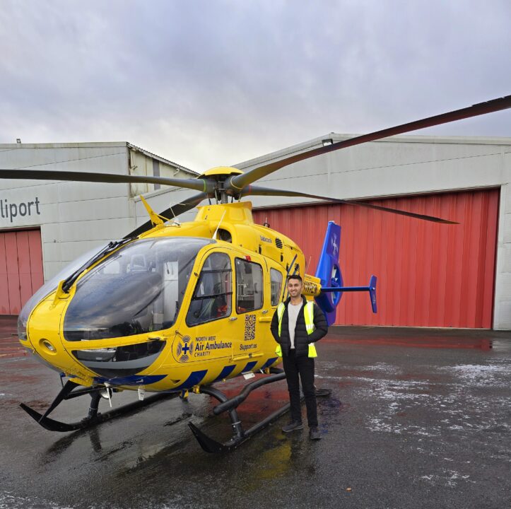 Ethan, wearing black jeans and a black jacket, stood on the helipad in front of the Charity's yellow and blue helicopter