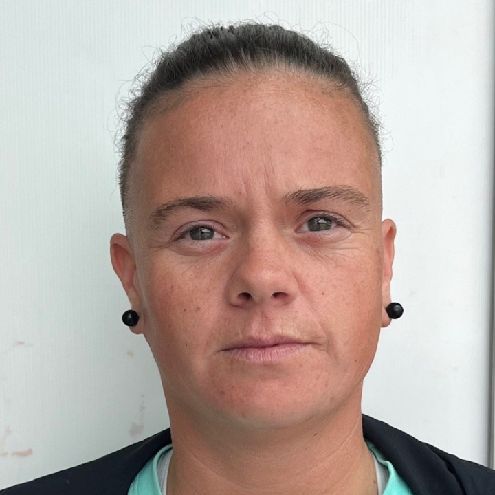 Headshot of a woman with light skin and tied-back hair, wearing small stud earrings, looking directly at the camera against a plain background