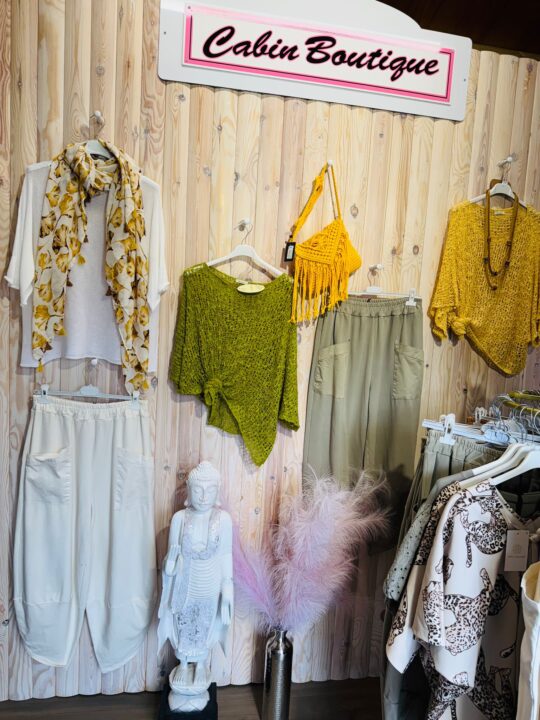 Clothing display inside Cabin Boutique featuring neutral and earthy-toned outfits. Items include cream trousers with a floral scarf, a green knit top, khaki trousers, a mustard knit top, and a yellow woven handbag. The display is styled against a light wooden wall with decorative pieces including a white Buddha statue and a vase with soft pink pampas grass