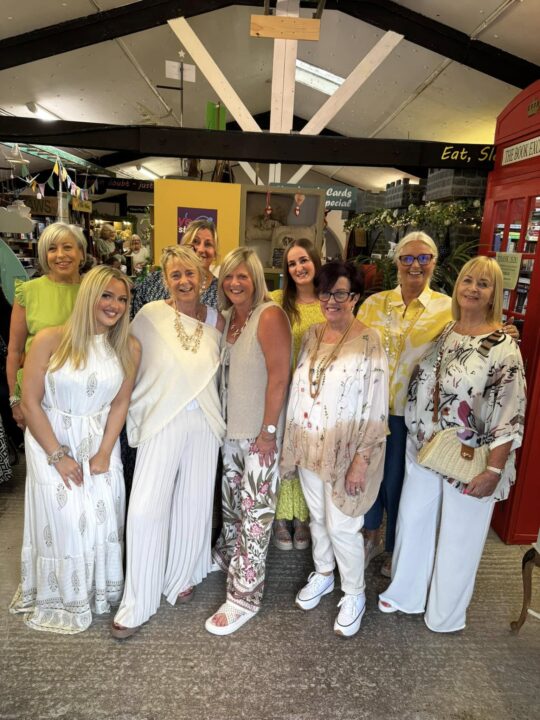 Group photo of eight women standing together indoors at Cabin Boutique. They are smiling and dressed in coordinated summer outfits in shades of white, cream, yellow, and floral patterns. The background shows boutique décor with bunting, signage, and a red British-style phone booth