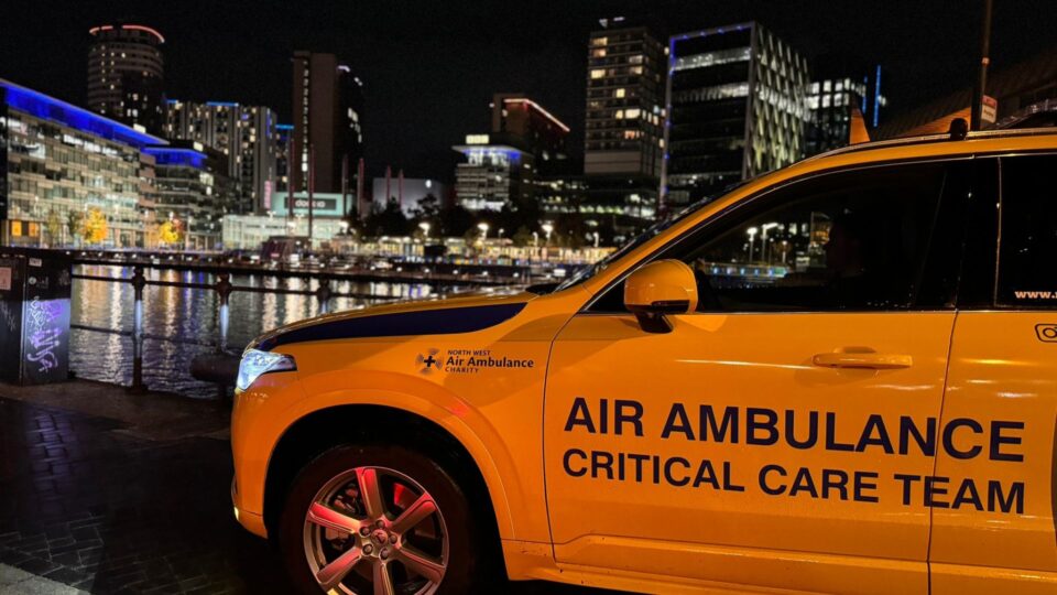 A yellow North West Air Ambulance critical care vehicle parked at night by the waterfront in a city, with tall modern buildings and colorful lights reflecting on the water in the background.