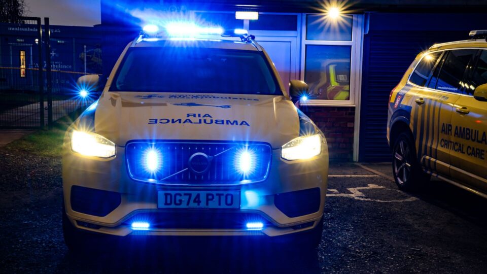 A yellow Volvo air ambulance critical care vehicle with bright blue emergency lights flashing at night, parked outside a building with "Air Ambulance" markings visible. Another response vehicle is partially visible on the right side.