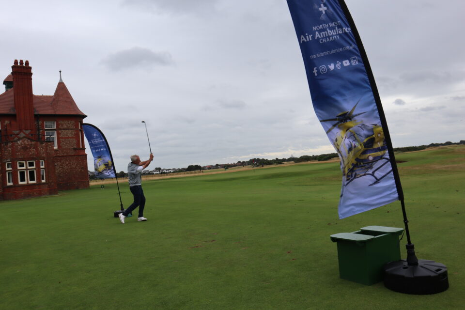 A golfer mid-swing on a green in front of a red-brick clubhouse, with a blue North West Air Ambulance Charity banner in the foreground featuring a yellow helicopter and social media logos