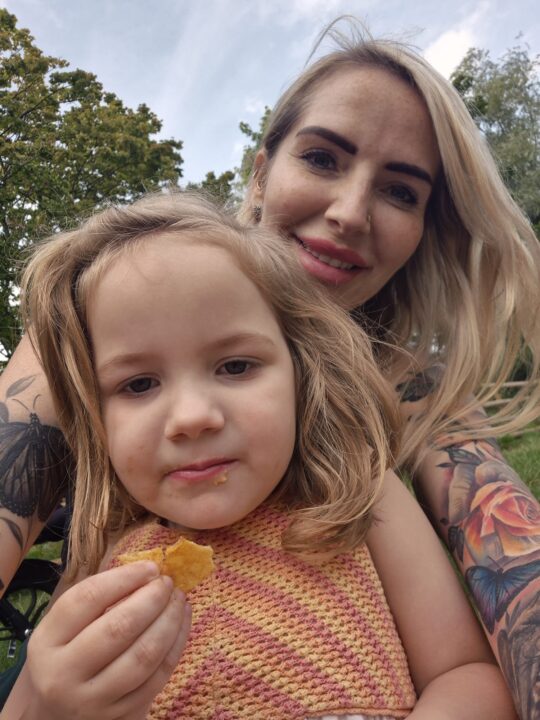 A woman with long blonde hair and tattoos smiles at the camera while sitting outdoors with a young girl. The child, wearing a knitted pink and yellow top, holds a piece of food and looks into the camera, with trees and sky in the background.