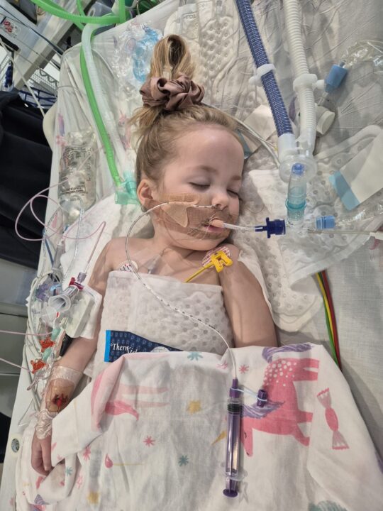 A young child lies in a hospital bed, asleep and connected to medical equipment. The child has bandages on their arm, monitoring stickers on their chest, and a breathing tube secured with tape, surrounded by wires and hospital bedding decorated with dinosaurs.
