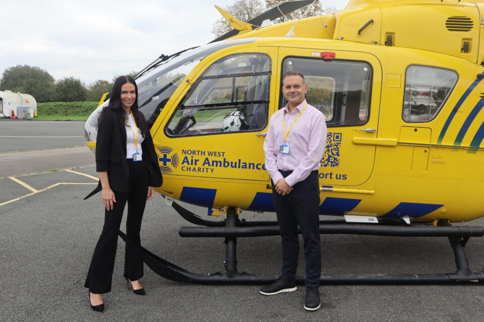 Two people standing in front of a yellow North West Air Ambulance Charity helicopter on a helipad, both wearing charity ID lanyards and smiling at the camera.