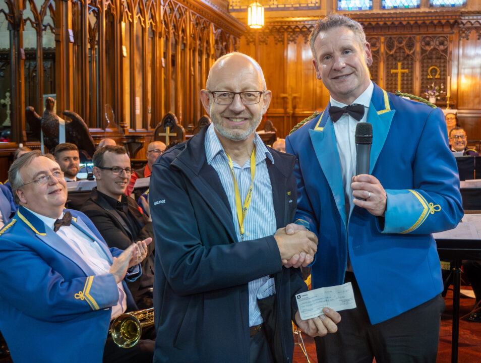 A band member in a blue uniform presents a cheque to a man wearing glasses and a dark jacket inside a church. Other band members sit behind them smiling and applauding