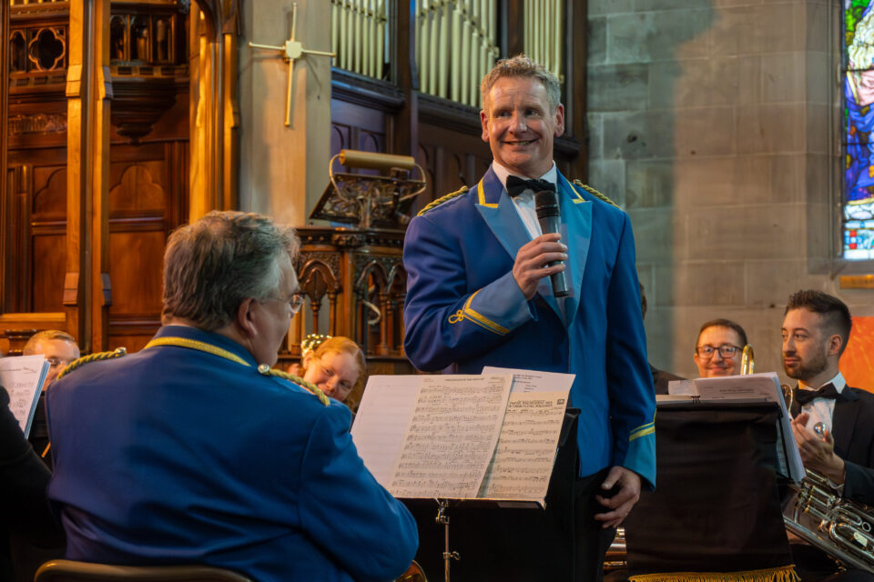 A musician in a bright blue band uniform smiles while holding a microphone, speaking to fellow band members during a performance in a church with ornate wooden panels and organ pipes in the background.