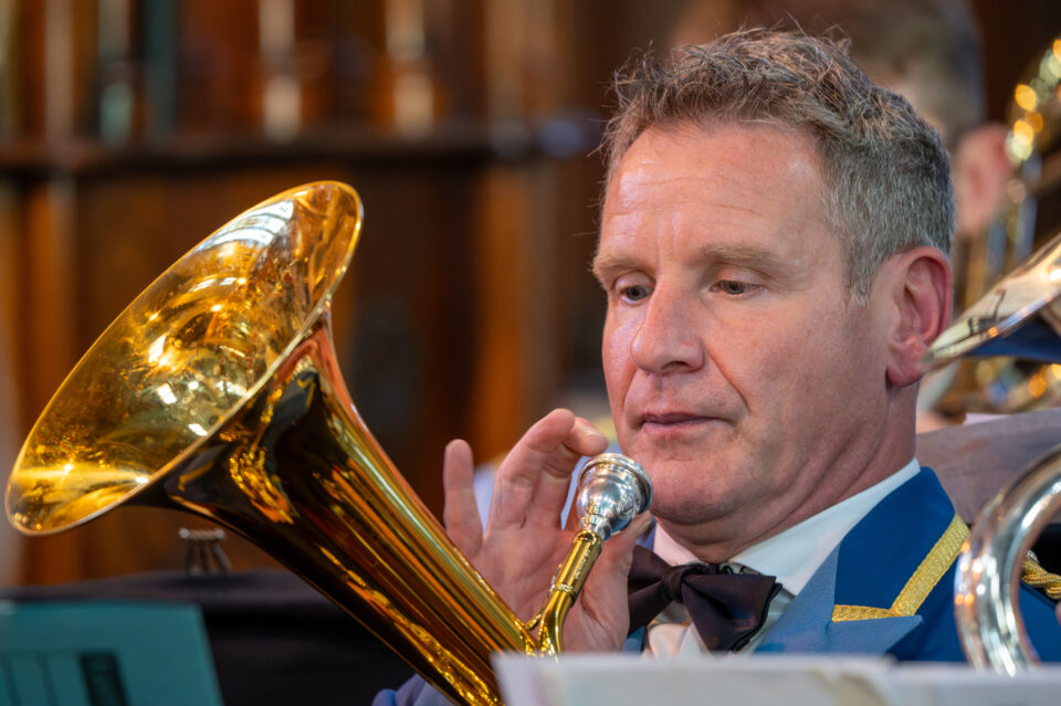 A close-up of a brass musician in a blue uniform focused on playing his instrument, with warm light reflecting off the polished surface of the horn