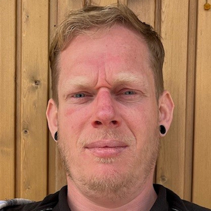 A close-up portrait of a person standing in front of a wooden wall, looking directly at the camera with a neutral expression.