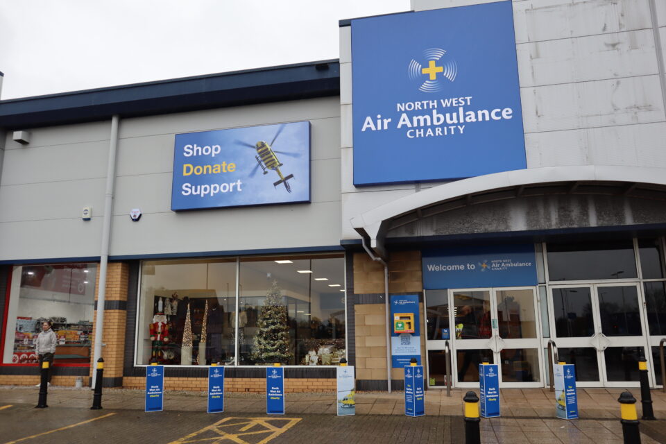 The exterior of a North West Air Ambulance Charity store, featuring large signs reading ‘Shop Donate Support’ and the charity logo. The shop window displays Christmas decorations, including a lit tree, and donation stands are positioned along the front walkway.
