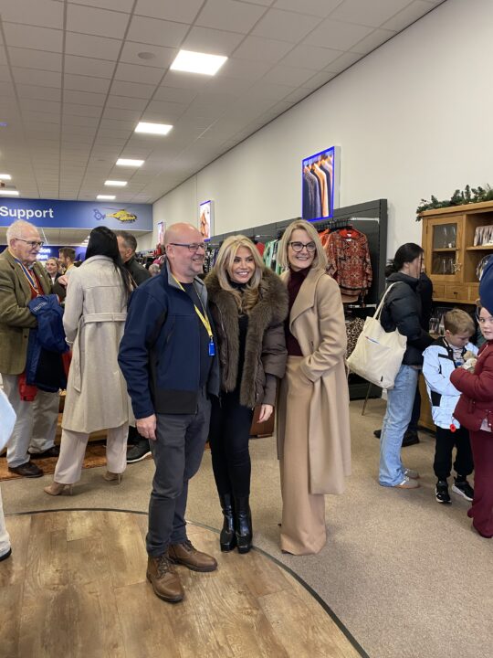 Three people pose together inside a busy North West Air Ambulance Charity store during an opening event. Shoppers and supporters fill the background as they stand in front of clothing displays and smile for the camera.