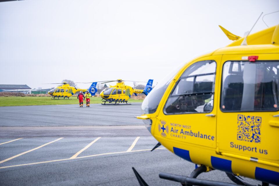 Three yellow North West Air Ambulance helicopters parked on a helipad, with two crew members in red and yellow uniforms walking toward one of the aircraft. The foreground shows a close-up of another helicopter featuring the charity's logo and a QR code. The scene takes place on an overcast day at an airfield.
