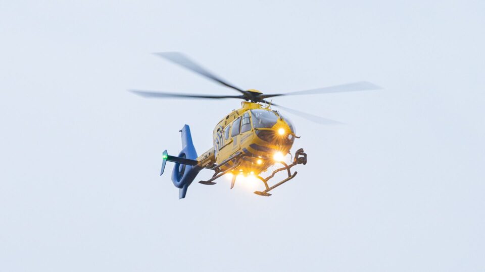 A yellow North West Air Ambulance Charity helicopter flying against a pale sky, with its landing lights on and rotor blades blurred in motion.