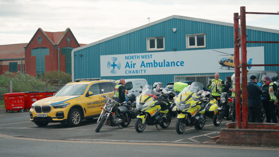 Motorcyclists gather outside the North West Air Ambulance Charity base, with several bikes parked alongside a yellow critical care response vehicle in front of the charity’s building and signage.