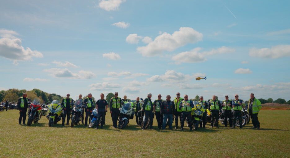 A large group of motorcyclists wearing high-visibility vests stand in a line with their bikes on a grassy field under a blue sky, with a North West Air Ambulance helicopter flying in the distance above them.