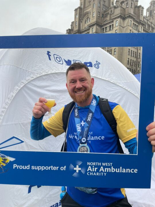 A man wearing a North West Air Ambulance Charity T-shirt and medal smiles while holding a drink and posing in a branded photo frame at a running event