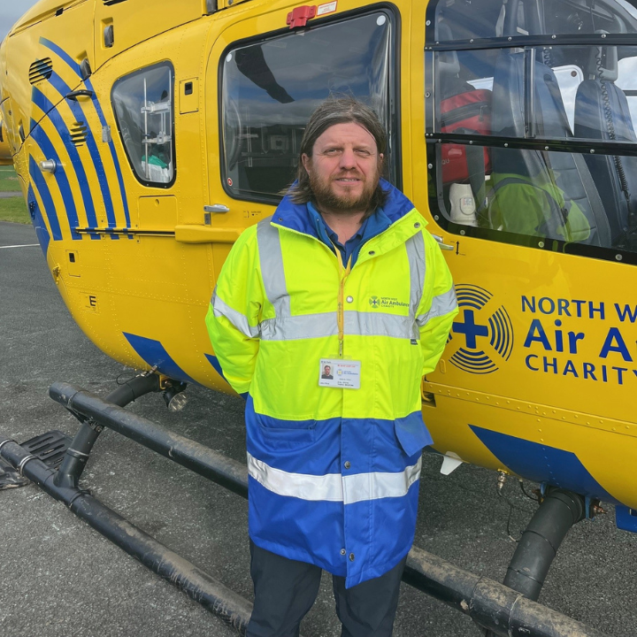 A man wearing a high-visibility North West Air Ambulance Charity jacket and ID badge stands in front of a yellow air ambulance helicopter.