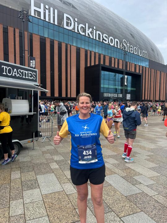 A woman wearing a North West Air Ambulance Charity running top and race number stands outside Hill Dickinson Stadium giving a thumbs-up among a crowd of runners