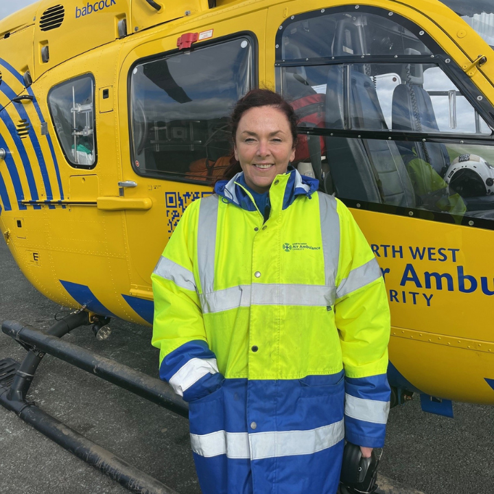 A woman wearing a high-visibility North West Air Ambulance Charity jacket stands smiling in front of a yellow air ambulance helicopter