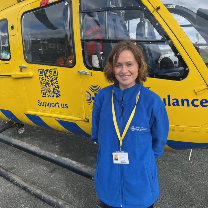A woman wearing a blue North West Air Ambulance Charity fleece and ID lanyard stands smiling beside a yellow air ambulance helicopter