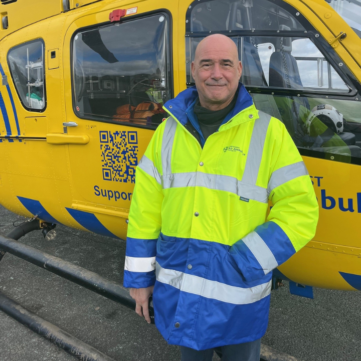 A man wearing a high-visibility North West Air Ambulance Charity jacket stands smiling beside a yellow air ambulance helicopter.