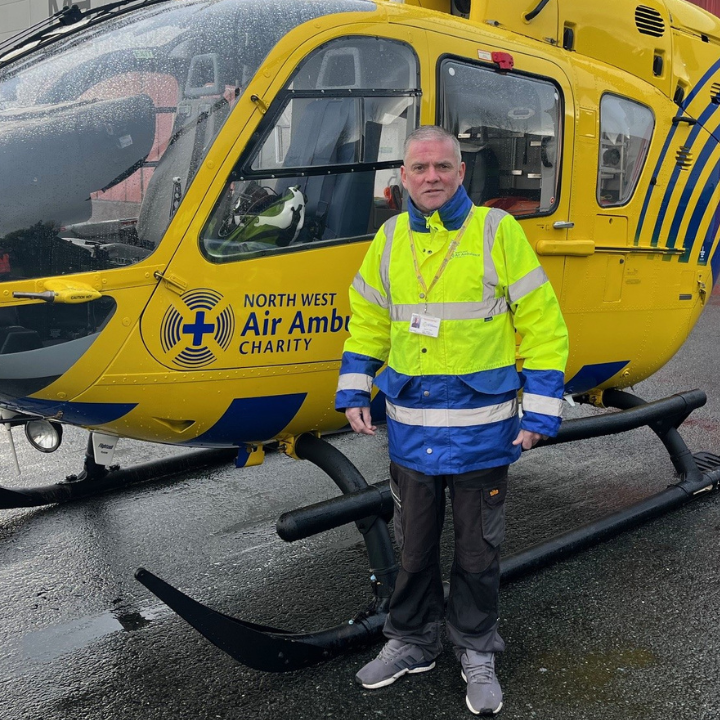 A man wearing a high-visibility North West Air Ambulance Charity jacket stands in front of a yellow air ambulance helicopter on a rainy day.