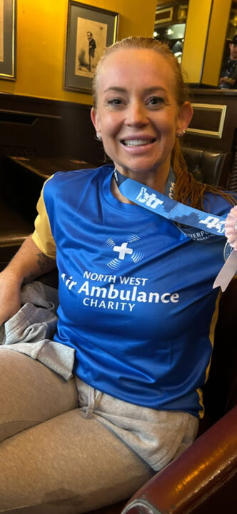 A woman wearing a blue North West Air Ambulance Charity T-shirt sits indoors smiling, with a Liverpool Marathon medal around her neck.
