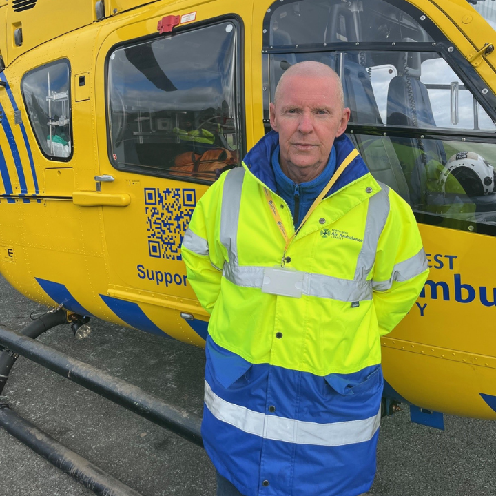 A man wearing a high-visibility North West Air Ambulance Charity jacket stands in front of a yellow air ambulance helicopter