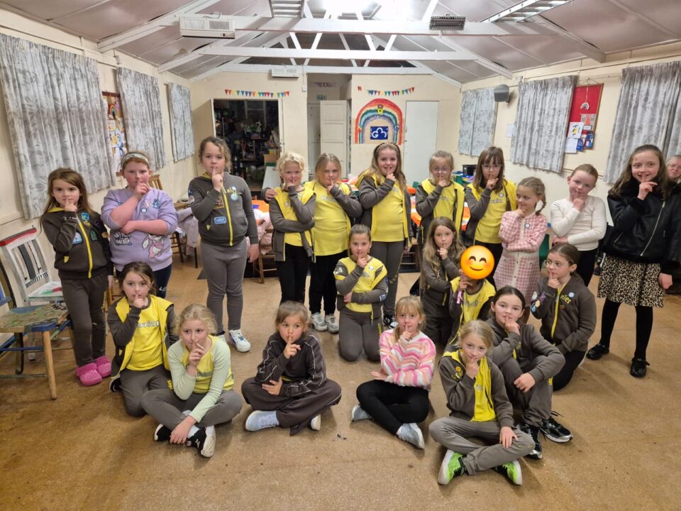A group of young girls in Brownies uniforms stand and sit together in a community hall, each holding a finger to their lips in a ‘shh’ pose. The room has tables, chairs, and colourful decorations including a rainbow on the wall