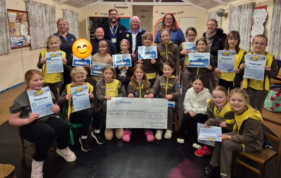 A group of Brownies and adult leaders pose together indoors, smiling and holding certificates, with two girls at the front holding a large cheque for £100 for North West Air Ambulance Charity.