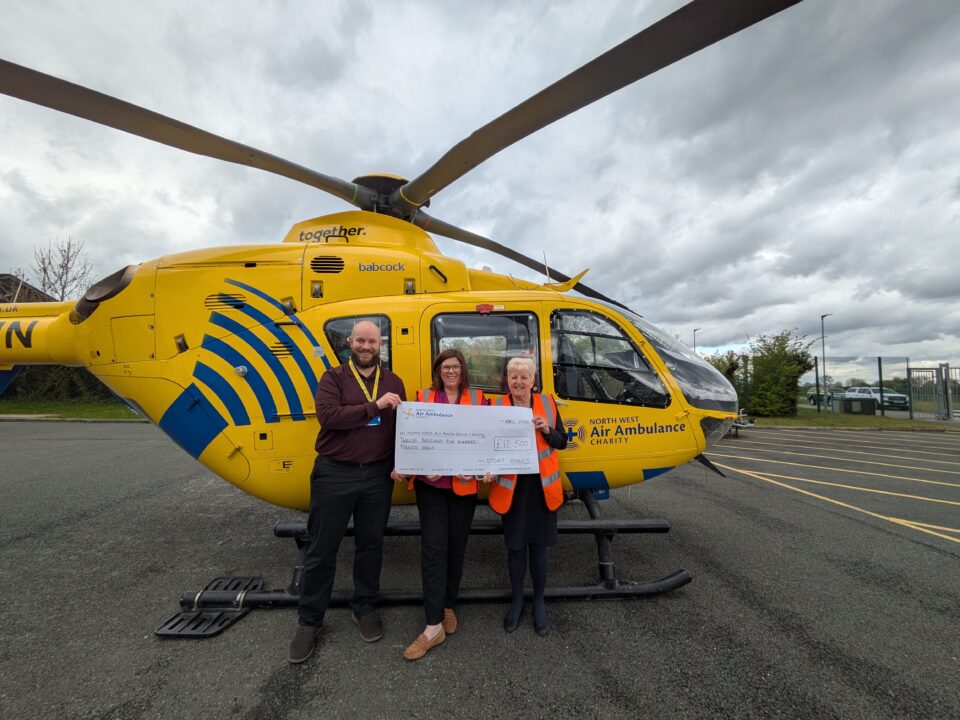 Three people stand in front of a yellow North West Air Ambulance helicopter, smiling and holding a large cheque for £12,500. The helicopter is parked on a tarmac area under a cloudy sky.