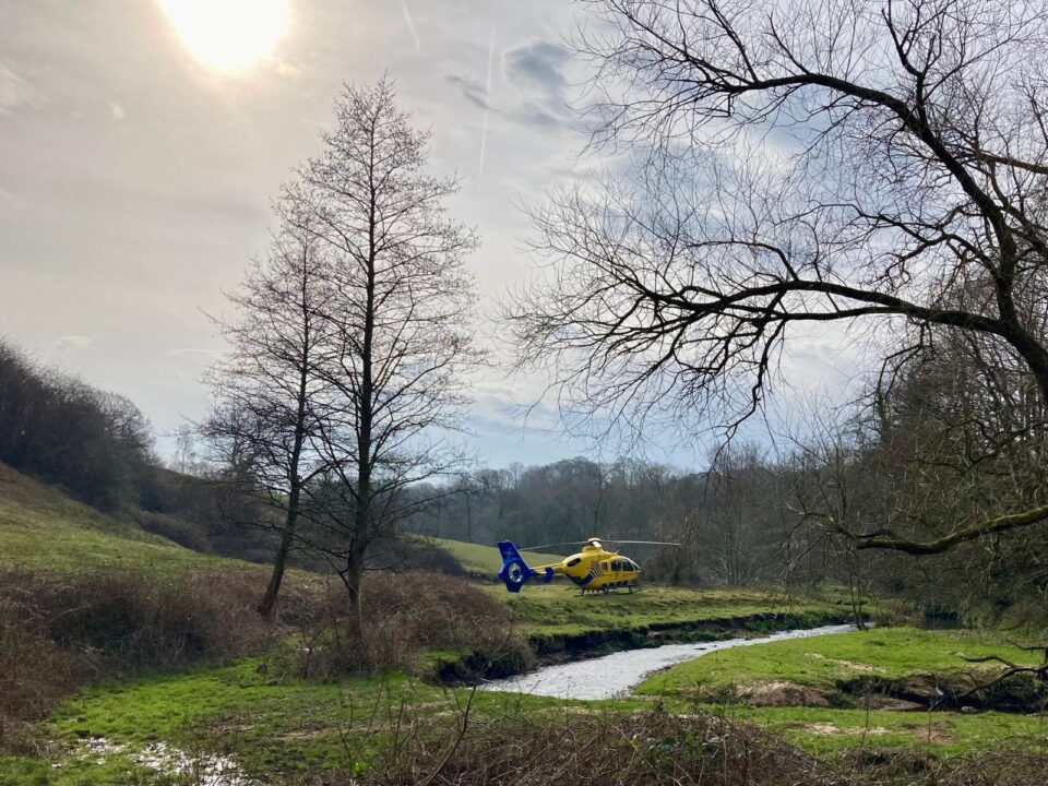 Viewed from a distance, a yellow and blue North West Air Ambulance helicopter is landed in a secluded rural landscape. A stream curves through the foreground, while leafless trees and grassy banks frame the scene. Sunlight breaks through a lightly clouded sky, highlighting the remote location of the emergency response landing site.