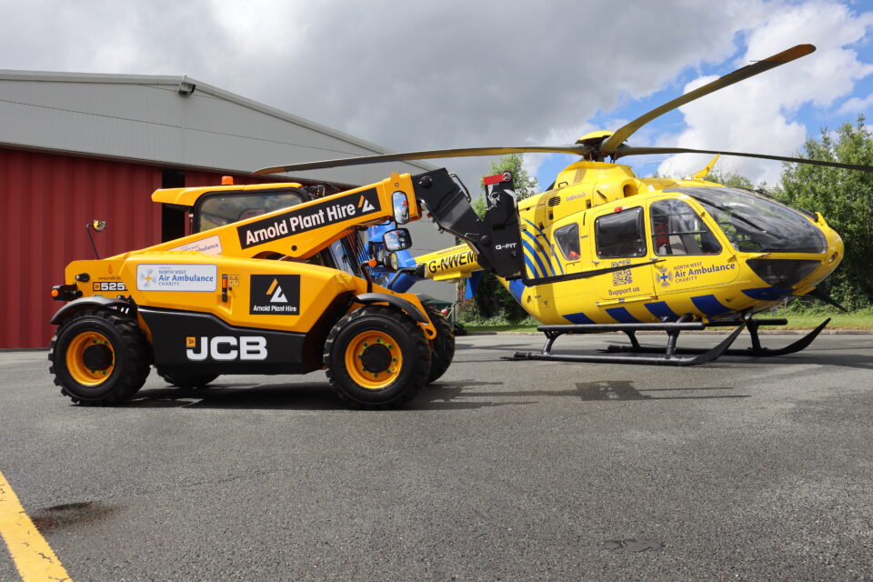 An Arnold Plant Hire JCB telehandler featuring North West Air Ambulance Charity branding stands next to one of the charity’s helicopters on the tarmac outside a hangar.