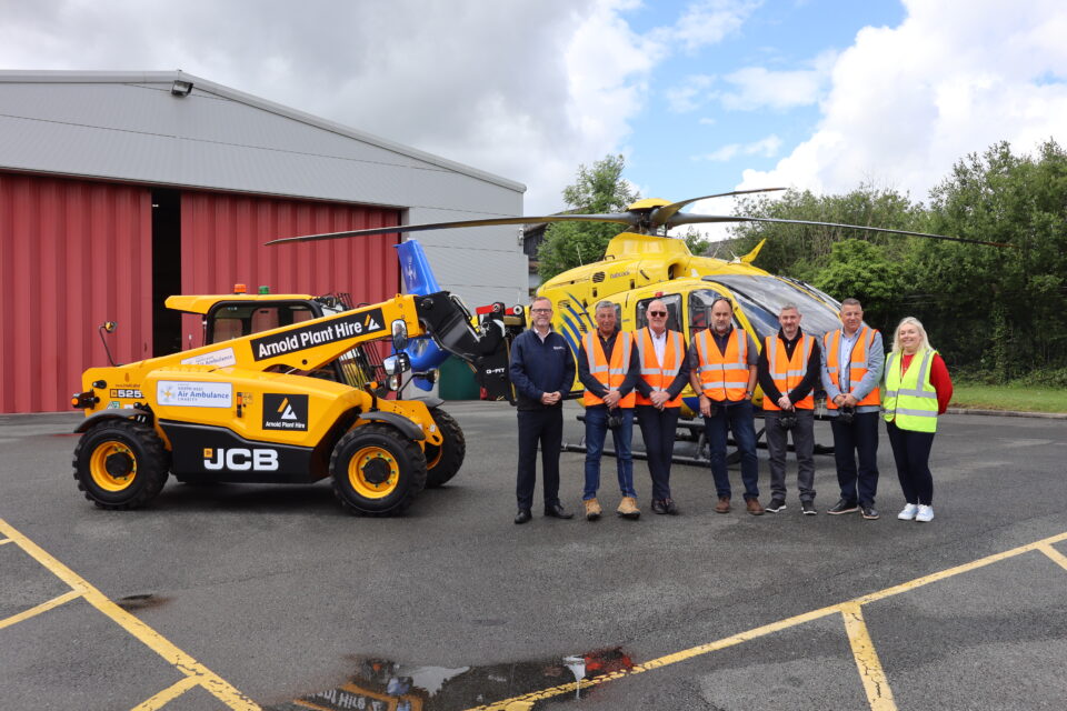 Staff from Arnold Plant Hire and the North West Air Ambulance Charity gather beside a branded JCB telehandler and air ambulance helicopter outside the charity’s base, celebrating their partnership.