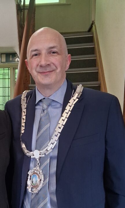 The Mayor of Bollington, wearing a ceremonial chain of office over a navy suit and striped tie, poses for a portrait inside a civic building.