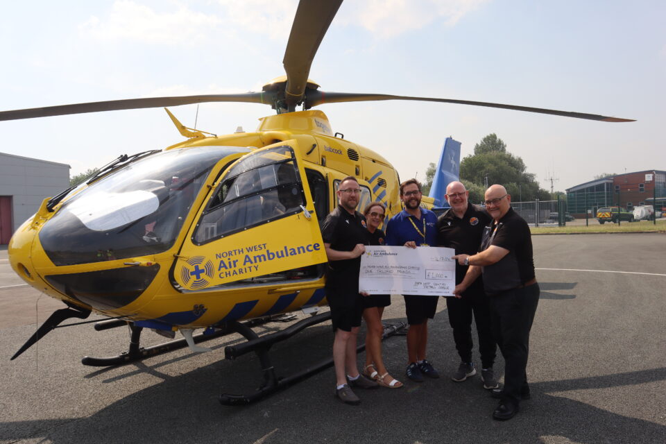 A group of five people stand beside a yellow North West Air Ambulance Charity helicopter on an airfield, holding a ceremonial cheque for £1,000. The helicopter's rotor blades are visible overhead, and airport buildings can be seen in the background.