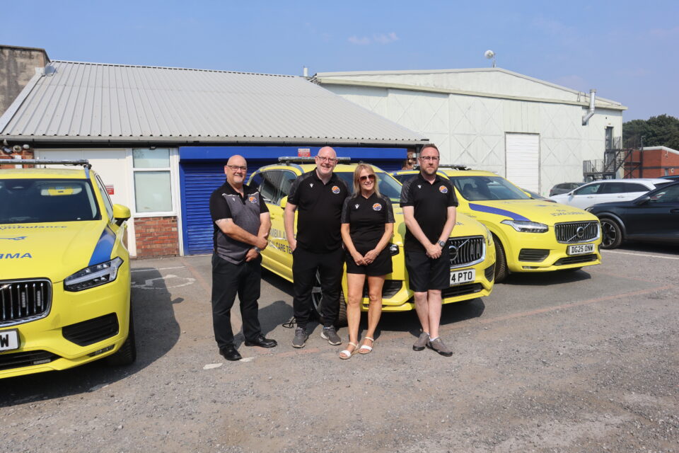 Four people stand in front of several yellow North West Air Ambulance Charity rapid response vehicles outside a service building on a sunny day. The branded emergency vehicles are parked behind the group in a yard area.