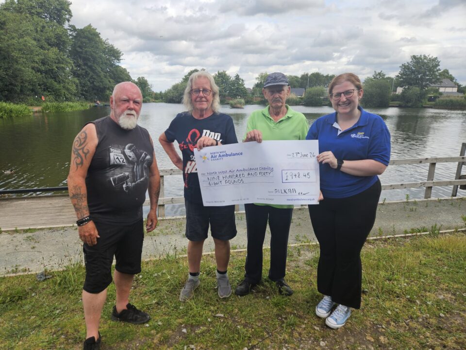 Four adults stand beside a lake holding an oversized ceremonial cheque made out to the North West Air Ambulance Charity for £948.45. The group poses outdoors near the same lakeside setting, with trees, houses, and a cloudy sky in the background.