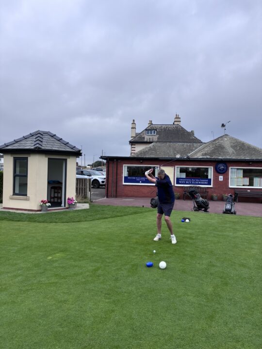 A golfer prepares to strike a ball on a well-maintained green at a golf club. A clubhouse, golf bags and parked cars can be seen behind the golfer under cloudy skies.