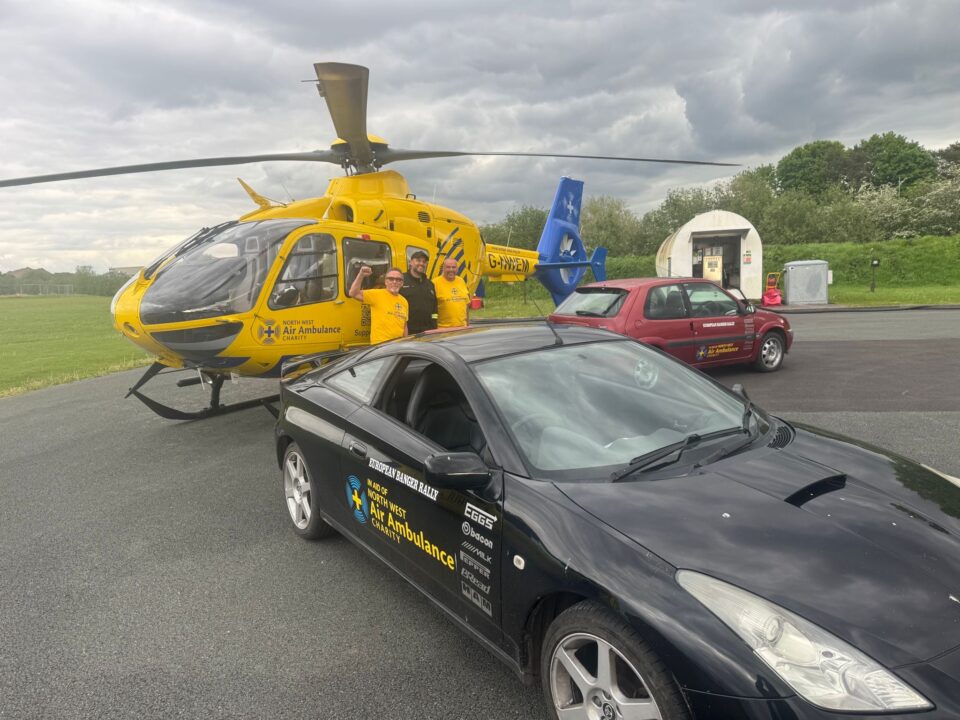 A yellow North West Air Ambulance Charity helicopter is parked on a tarmac area at an airfield next to two vehicles displaying the charity's branding. Three people in yellow charity T-shirts stand beside the aircraft, with grassy fields and airfield buildings visible in the background beneath dramatic grey clouds.