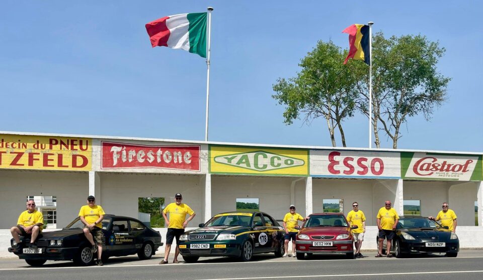 A group of charity supporters wearing yellow North West Air Ambulance Charity T-shirts pose alongside a collection of cars outside a historic-looking motorsport pit building. Vintage advertising signs for Firestone, Yacco, Esso and Castrol are displayed on the building, while Italian and Belgian flags fly above against a clear blue sky.